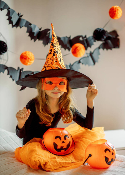 Маленькая девочка в костюме ведьмы с тыквенной конфеткой на Хэллоуин дома. Хаус пати Хэллоуин. Праздничный декор  - Фото, изображение