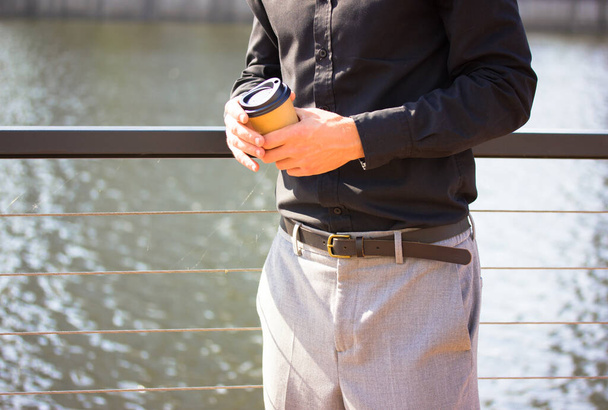 A businessman in a black shirt, gray pants stays on a bridge, pier, drinking cappuccino latte espresso from a kraft paper cup. Coffee to go. Rest, chill by a sea, river bank, lake. Man relaxes outdoor - Фото, зображення
