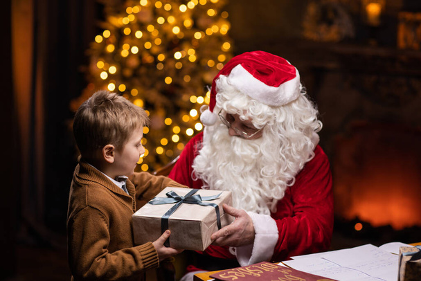 Santa Claus and little boy. Cheerful Santa is working while sitting at the table. Fireplace and Christmas Tree in the background. Christmas concept. - 写真・画像