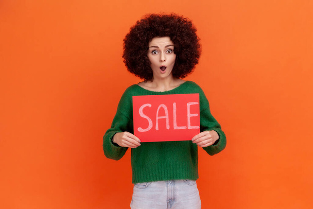 Shocked woman with Afro hairstyle in green casual style sweater holding card with sale inscription, standing with open mouth, astonishing discounts. Indoor studio shot isolated on orange background. - Zdjęcie, obraz