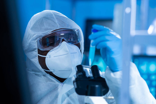 African-american doctor in protective suit is doing a science experiments and developing vaccine in a modern lab. Laboratory assistant workplace. The concept of science and medicine. - Фото, изображение