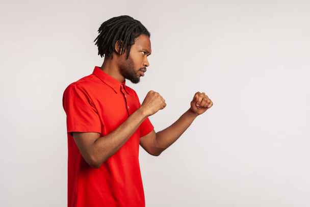 Profile portrait angry of man with dreadlocks wearing red casual style T-shirt, punching, boxing with clenched fists, being ready fighting. Indoor studio shot isolated on gray background. - Foto, Bild