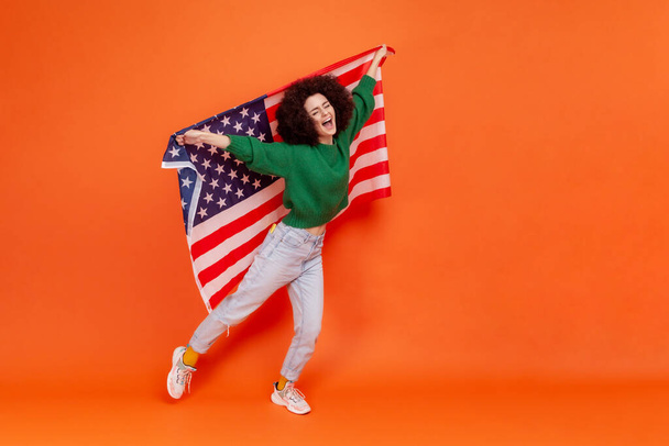 Plná délka fotografie ženy s Afro účes na sobě zelené ležérní styl svetr zvedl ruce, drží americkou vlajku, slaví národní svátek. Vnitřní studio záběr izolované na oranžovém pozadí. - Fotografie, Obrázek