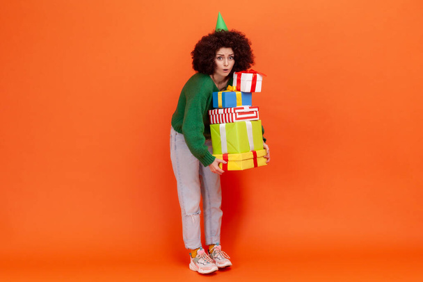 Full length portrait of woman with Afro hairstyle wearing green sweater holding lots of heavy presents, looking at camera with astonished expression. Indoor studio shot isolated on orange background. - Fotó, kép