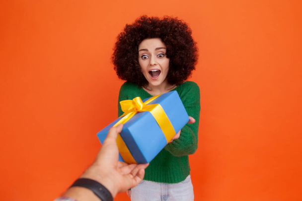 Portrait of happy excited woman with Afro hairstyle wearing green casual style sweater getting blue wrapped present box from unknown person. Indoor studio shot isolated on orange background. - Foto, Imagen