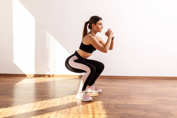 Full length portrait of woman doing split or lunge squat exercise at home or fitness gym, wearing black sports top and tights. Indoor studio shot illuminated by sunlight from window. - Fotó, kép