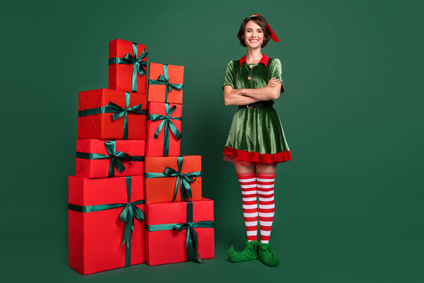 Photo of pretty confident young woman dressed elf costume standing gift boxes pile arms crossed smiling isolated green color background - Foto, afbeelding