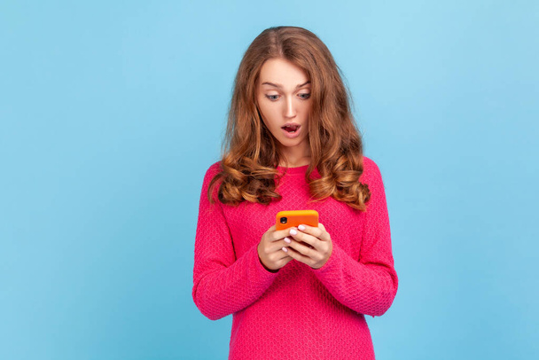 Portrait of amazed woman wearing pink pullover, looking at cellphone with surprised expression, reading shocking news using mobile phone. Indoor studio shot isolated on blue background. - Photo, image