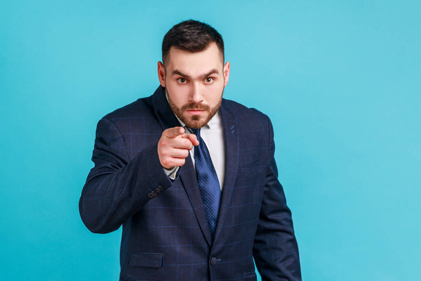 Serious bearded man wearing official style suit pointing finger, indicating direction to camera and looking annoyed, making choice. Indoor studio shot isolated on blue background. - Photo, image
