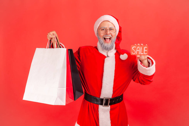 Portrait of happy satisfied elderly man with gray beard wearing santa claus costume holding shopping bags and sale cards, mall discounts. Indoor studio shot isolated on red background. - Zdjęcie, obraz