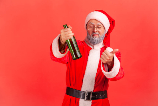 Funny elderly man with gray beard wearing santa claus costume standing with beer bottle, being drunk, standing with closed eyes and showing thumb up. Indoor studio shot isolated on red background. - Photo, image