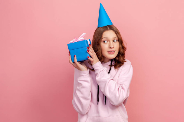 Portrait of curious curly haired teenage girl in hoodie with party hat holding wrapped gift box, trying to understand what inside. Indoor studio shot isolated on pink background - Fotografie, Obrázek