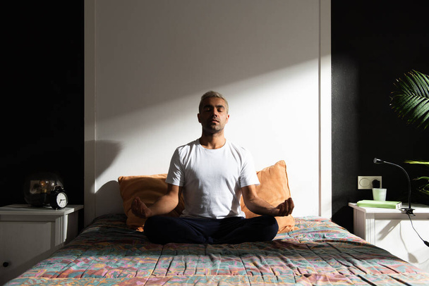 太陽の光で照らされた寝室で瞑想するヒスパニック系の男性。健康的な朝のルーチンの概念 - 写真・画像