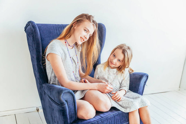 Two happy kids sitting on cozy blue chair relaxing playing in white living room indoors. Little girl playing with teenage girl showing her love care. Sisters having fun at home - Photo, Image