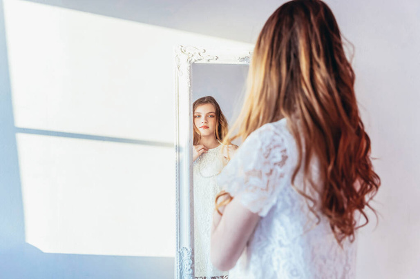 Beauty makeup morning rutine love yourself concept. Young teenage girl looking at reflection in mirror. Young positive woman wearing white dress posing in bright light room against white wall - Foto, afbeelding