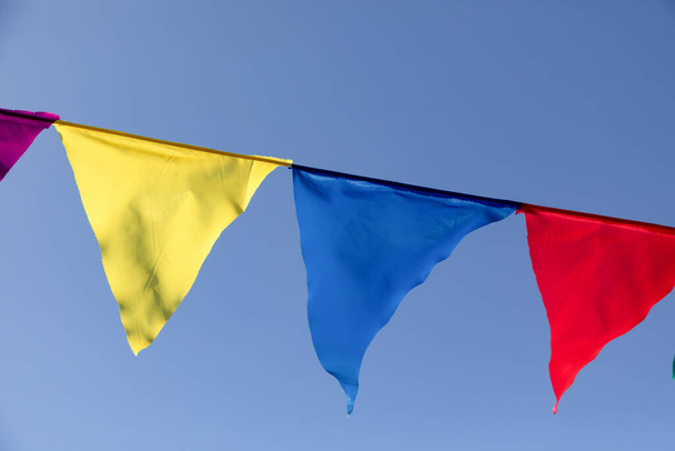 Κίτρινες, μπλε και κόκκινες τριγωνικές σημαίες στο φόντο ενός καθαρού καλοκαιρινού ουρανού. Πάρτι εορτασμού εξωτερικού χώρου. Γιορτινή διάθεση - Φωτογραφία, εικόνα