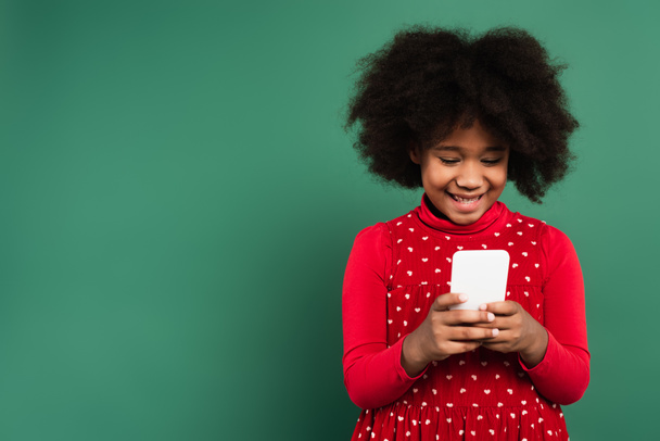 Uśmiechnięta Afroamerykanka w czerwonej sukience za pomocą telefonu komórkowego na zielonym tle  - Zdjęcie, obraz