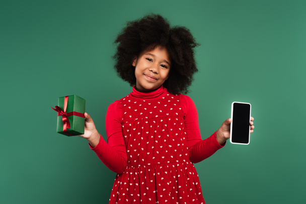 Smiling african american child in red dress holding present and smartphone with blank screen isolated on green  - Photo, Image