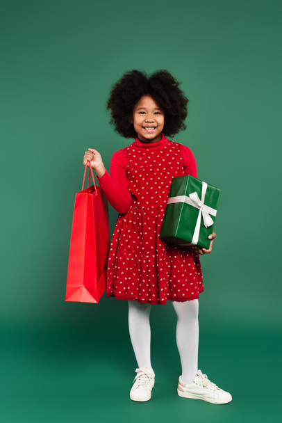 Позитивна афро-американська дівчинка, яка тримає подарунок і червоний пакет на зеленому фоні  - Фото, зображення