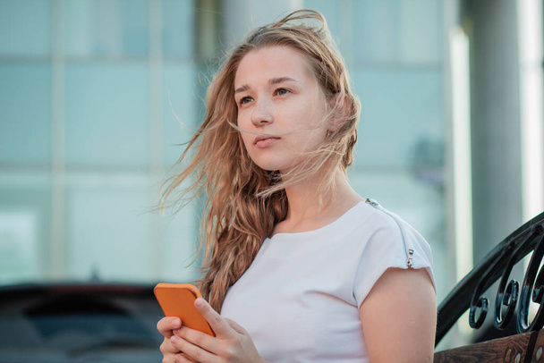 Beyaz elbiseli akıllı bir telefonla bankta oturan genç bir kadın. İş binasının yakınında telefonla konuşan bir kız. - Fotoğraf, Görsel