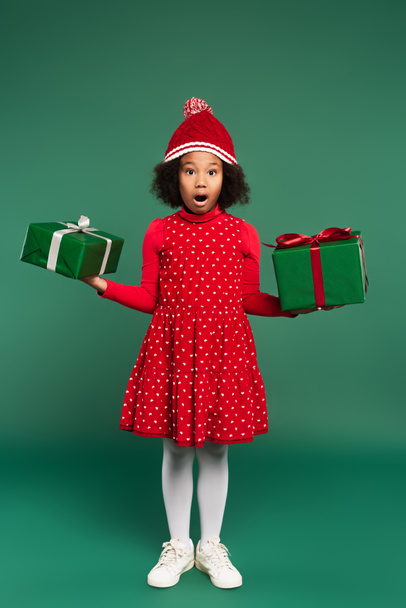 Verbaasd Afrikaans Amerikaans kind in jurk en hoed met geschenken op groene achtergrond  - Foto, afbeelding