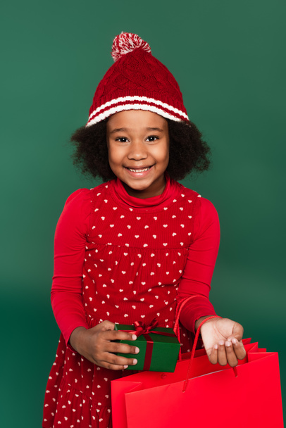 Enfant afro-américain joyeux dans un chapeau tenant un sac à provisions et présent isolé sur vert  - Photo, image