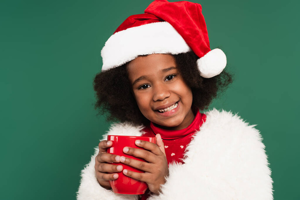 Positive african american kid in santa hat and fluffy jacket holding red cup isolated on green  - Photo, Image