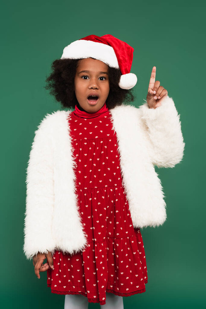 Aufgeregtes afrikanisch-amerikanisches Mädchen in flauschiger Jacke und Weihnachtsmütze mit Idee auf grün  - Foto, Bild