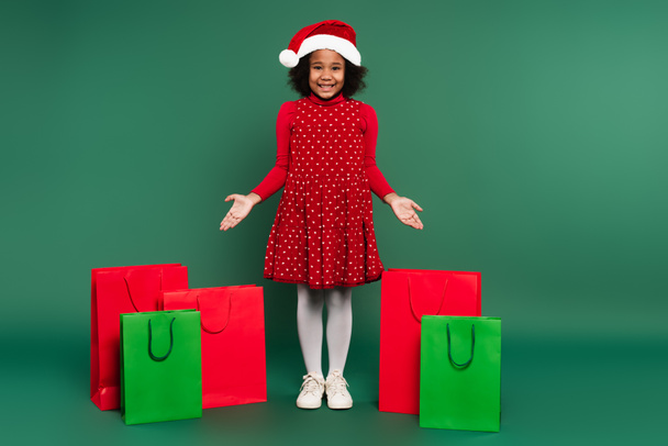 Lächelndes afrikanisch-amerikanisches Kind in Kleid und Weihnachtsmütze zeigt auf Einkaufstüten auf grünem Hintergrund  - Foto, Bild