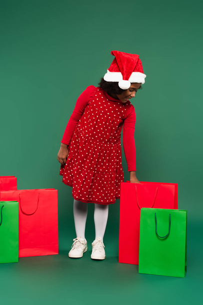 Side view of positive african american girl in santa hat and red dress looking at shopping bag on green background  - Valokuva, kuva