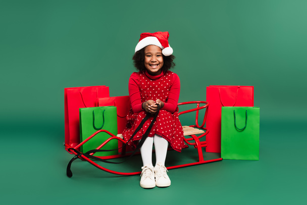 Smiling african american kid in santa hat sitting on sleigh near shopping bags on green background  - Fotó, kép