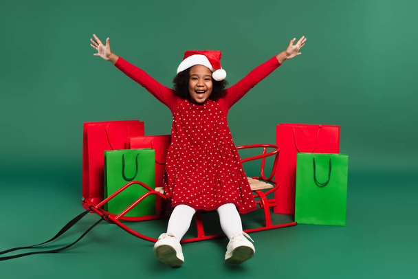 Aufgeregtes afrikanisch-amerikanisches Kind in Kleid und Weihnachtsmütze auf Schlitten neben Einkaufstaschen auf grünem Hintergrund  - Foto, Bild