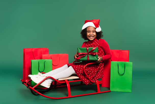 African american girl in santa hat holding present while sitting on sleigh near shopping bags on green background  - Fotoğraf, Görsel