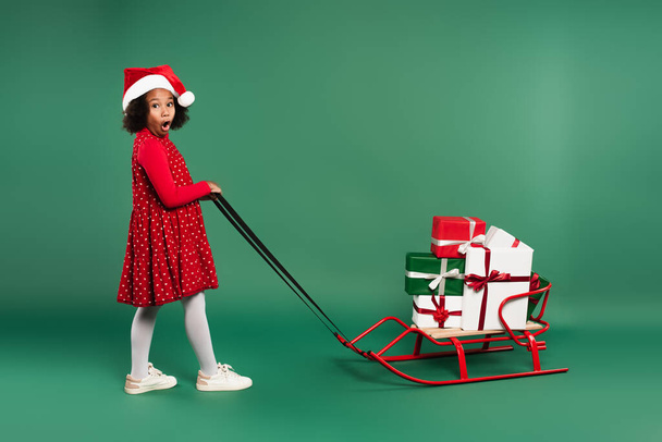 Amazed african american girl in santa hat pulling sleigh with gifts on green background  - Photo, Image