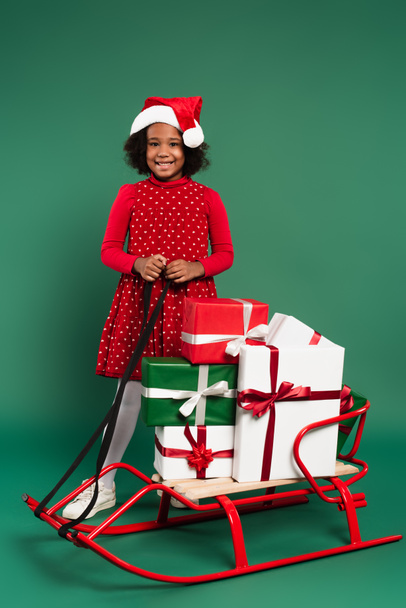 Allegro bambino africano americano in cappello di Babbo Natale in piedi vicino slitta con regali su sfondo verde  - Foto, immagini