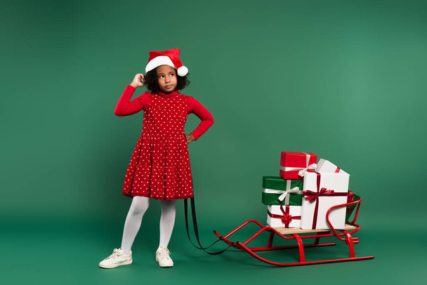 Pensive african american kid in red santa hat standing near gifts on sleigh on green background  - Photo, Image