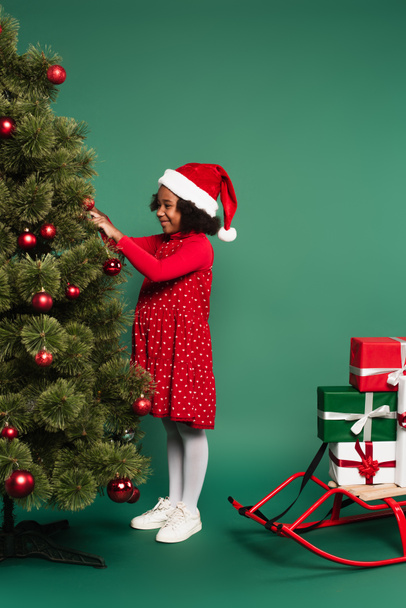 Afrikaans amerikaans kind in santa hoed versieren kerstboom in de buurt slee met geschenken op groene achtergrond  - Foto, afbeelding