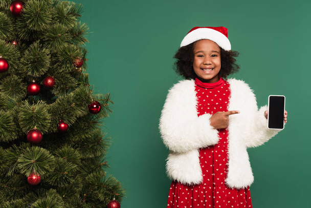 Positive african american girl in santa hat and fluffy jacket pointing at smartphone near decorated christmas tree isolated on green - Photo, Image