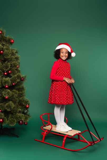Smiling african american child in dress and santa hat standing on sleigh near christmas tree on green background  - Photo, Image