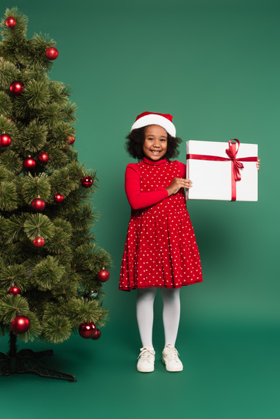 Noel Baba şapkalı mutlu Afrikalı Amerikalı kız yeşil arka planda dekore edilmiş Noel ağacının yanında hediye kutusu tutuyor.  - Fotoğraf, Görsel