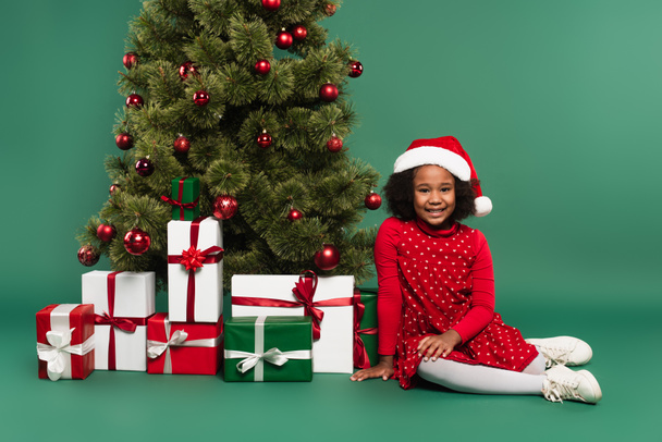 African American κορίτσι στο καπέλο Σάντα κάθεται κοντά σε δώρα και χριστουγεννιάτικο δέντρο σε πράσινο φόντο  - Φωτογραφία, εικόνα