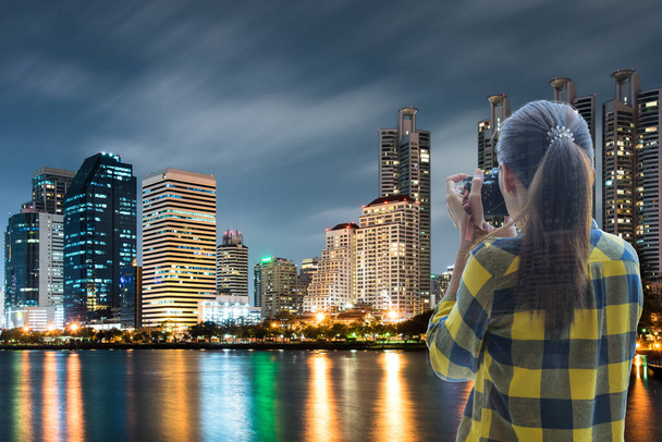 Doble exposición de una mujer vestida con una camisa amarilla a cuadros y sosteniendo la cámara para tomar una foto con fondo de paisaje urbano nocturno de fondo. - Foto, imagen