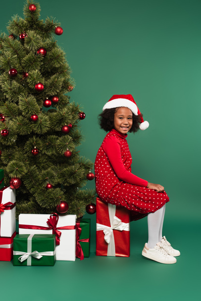 gelukkig afrikaans amerikaans kind in santa hoed zitten op aanwezig in de buurt van kerst boom op groene achtergrond  - Foto, afbeelding