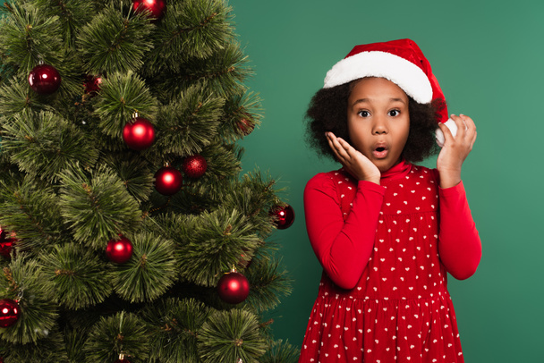 Verbazingwekkend Afrikaans amerikaans kind in santa hoed kijken naar camera in de buurt van kerst boom op groene achtergrond  - Foto, afbeelding