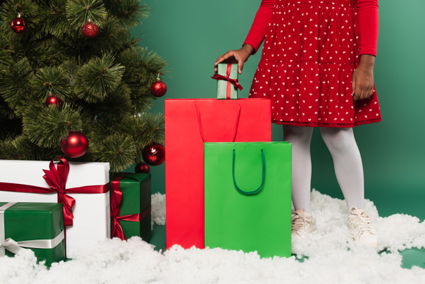 Ausgeschnittene Ansicht von afrikanisch-amerikanischen Kind setzen Geschenk in Einkaufstasche auf dekorativen Schnee in der Nähe von Weihnachtsbaum auf grünem Hintergrund  - Foto, Bild