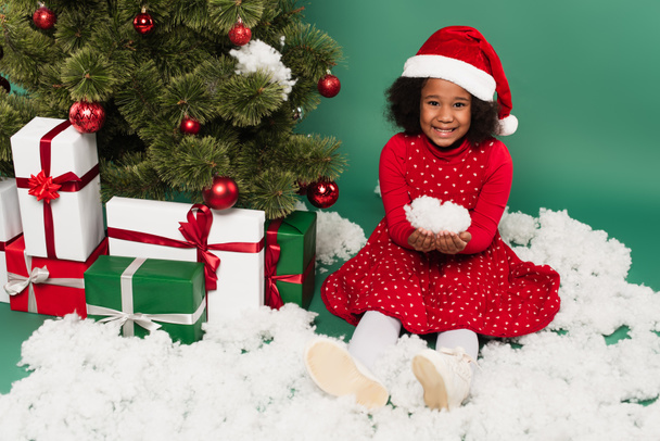 Enfant afro-américain en chapeau de Père Noël tenant la neige décorative près des cadeaux et arbre de Noël sur fond vert  - Photo, image
