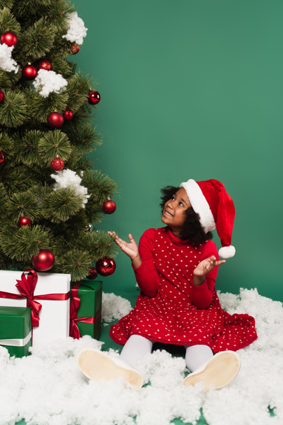 Africké americké dítě v Santa klobouk ukazuje na vánoční strom s dárky na dekorativní sníh na zeleném pozadí  - Fotografie, Obrázek