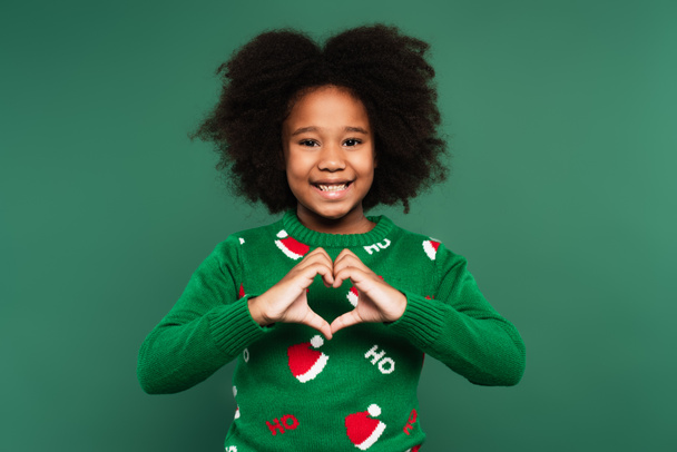 Criança americana africana alegre em suéter quente mostrando sinal de amor isolado no verde - Foto, Imagem