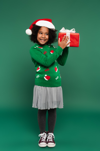 Cheerful african american girl in santa hat and sweater holding present on green background  - Photo, Image