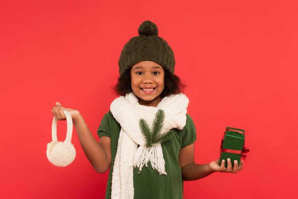 African american kid in hat and fir branch on scarf holding ear muffs and present isolated on red - Photo, Image